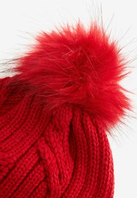 Bonnet tricoté rouge avec un motif de câble texturé, doté d'un grand pompon rouge duveteux sur le dessus. La laine douce et épaisse renforce la chaleur.