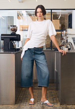 Femeie în top alb și pantaloni culotte din denim cu picioare largi stă între o mașină de cafea și un espressor într-un cadru modern de cafenea.