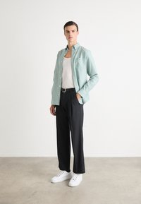Camicia verde chiaro con bottoni e tasca, indossata sopra una canottiera bianca. Pantaloni neri e sneakers bianche completano l'outfit. Sfondo semplice.