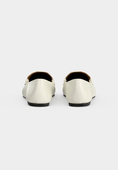 Ζευγάρι λευκά δερμάτινα loafers χωρίς κορδόνια, φαίνονται από πίσω, δείχνοντας χαμηλές μαύρες σόλες και μαλακή εσωτερική επένδυση σε μονόχρωμο φόντο.