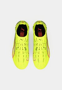 Tacos de fútbol de color amarillo brillante con una textura acanalada, con un logo negro de PUMA y detalles en rojo. Sistema de cordones visible en la parte frontal.