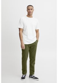 Mies seisoo yhdellä kädellä taskussa, pukeutuneena valkoiseen t-paitaan, oliivinvihreisiin housuihin ja mustiin lenkkareihin valkoista taustaa vasten.