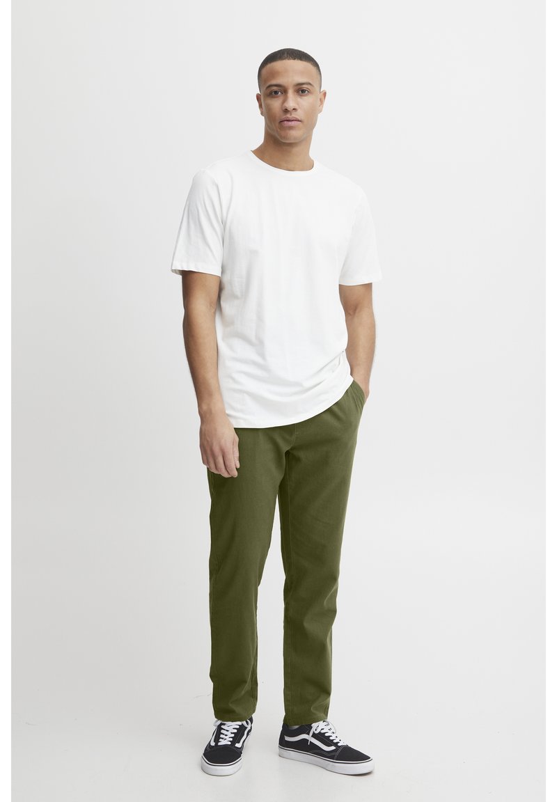 Mies seisoo yhdellä kädellä taskussa, pukeutuneena valkoiseen t-paitaan, oliivinvihreisiin housuihin ja mustiin lenkkareihin valkoista taustaa vasten.