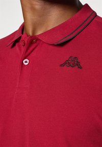 Červené polo tričko vyrobené z textúrovaného materiálu, s čiernym vyšitým logom, dvojitým zapínaním na gombíky a prúžkovaným detailom goliera.