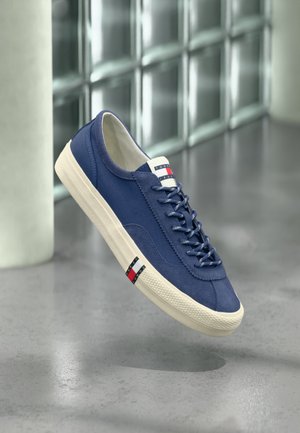 Basket en toile bleu marine avec semelle et lacets blancs, marquée du logo Tommy Hilfiger sur la languette et le côté, flottant au-dessus d'un sol poli.