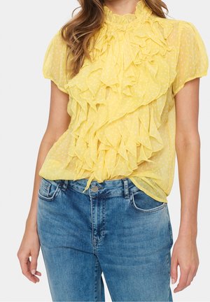 Nainen, jolla on päällään keltainen lyhythihainen blus, jossa on valkoisia pilkkuja ja röyhelöitä, yhdistettynä sinisiin farkkuihin.