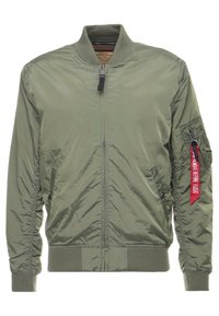 Blouson bomber vert olive avec fermeture éclair devant, col, poignets et ourlet côtelés, et une étiquette rouge « "Remove Before Flight" » sur la poche de la manche.