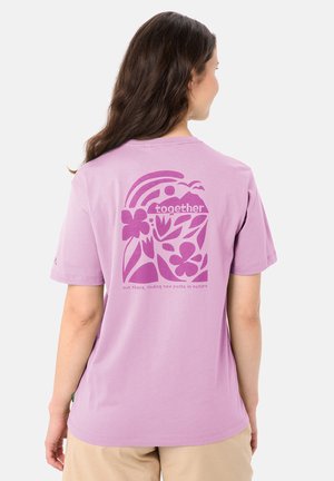 Donna con lunghi capelli castani che indossa una maglietta viola chiaro con grafica floreale e naturale e il testo "insieme" sul retro.