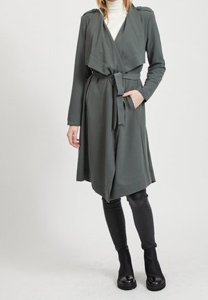 Femme portant un trench-coat ceinturé gris foncé par-dessus un col roulé blanc, leggings noirs et bottines noires, debout devant un fond uni.
