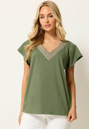 Blonde vrouw die een olijfgroene top met korte mouwen en geborduurde V-hals draagt en witte broek, staand tegen een egale achtergrond.