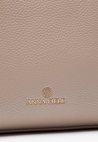 Beige texturerat läder med subtil kornstruktur, med en guld "ANNA FIELD" logotyp i bottenmitten.