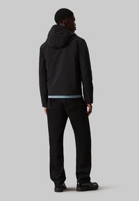 Giacca nera impermeabile con cappuccio, caratterizzata da una texture liscia e una silhouette aderente, abbinata a jeans neri e calzature nere.
