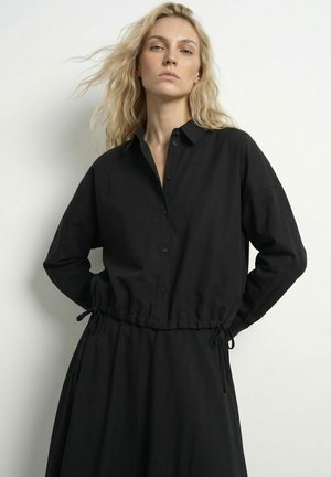 Femme blonde aux cheveux ondulés et détachés portant une robe noire à manches longues boutonnée avec un cordon à la taille, debout devant un fond clair uni.