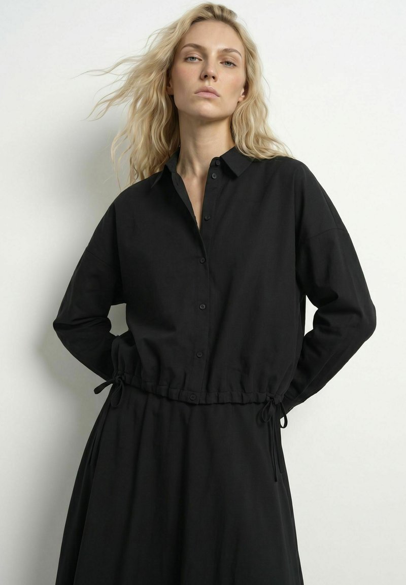 Femme blonde aux cheveux ondulés et détachés portant une robe noire à manches longues boutonnée avec un cordon à la taille, debout devant un fond clair uni.