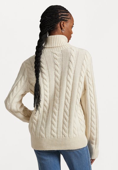 Dikke kabelgebreide trui in off-white met een hoge kraag. Kenmerkt zich door geribbelde manchetten en zoom. Gekomd met blauwe spijkerbroek, toont textuur en patroon.