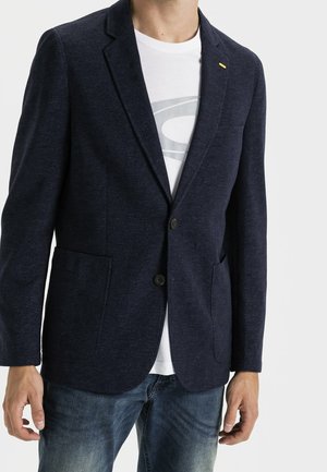 Hombre que lleva una chaqueta azul marino oscuro con dos botones sobre una camiseta blanca con diseño gráfico y vaqueros azules, de pie frente a un fondo liso.