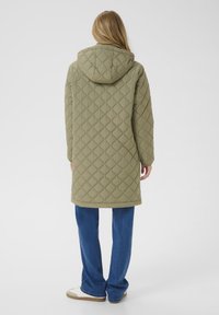 Kvinde med langt blond hår iført en kviltet olivengrøn frakke med hætte, blå jeans og hvide sneakers, vendt væk mod en ensfarvet baggrund.