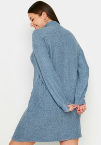 Abito in maglia blu con maniche lunghe, scollatura alta e vestibilità rilassata. Il tessuto sembra morbido e testa di pizzo, arrivando a metà coscia.