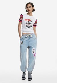 Witte cropped t-shirt met grafische print van een karakter en rode accenten, gecombineerd met lichtblauwe relaxed-fit jeans met bloemenpatches.