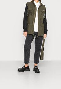 Olive green mixed-material jacket with black sleeves, multiple pockets, and detail patches. Paired with dark gray jeans and chunky black loafers.