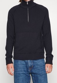 Svart stickad tröja med hög krage, med en kvartdragkedja, raglanärmar och en framficka i kängurustil. Slät textur.