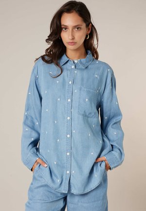Femme aux cheveux foncés portant une chemise en jean bleu clair à boutons avec de petits motifs de cœurs blancs, les mains dans les poches du pantalon assorti, fond neutre.