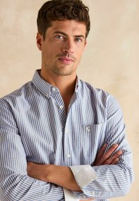 Koszula w paski, zapinana na guziki, w niebiesko-białym kolorze, wykonana z bawełny, z kołnierzykiem w szpic i kieszonką na piersi. Manżetki mają białe akcenty.