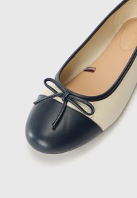 Gros plan sur une ballerine beige et marine avec un nœud marine sur l'orteil et un petit détail en tissu rouge, blanc et bleu à l'intérieur.