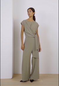 Khaki jumpsuit with a wrapped design, short sleeves, wide legs, and a belted waist. Smooth fabric with minimal stitching detail.
