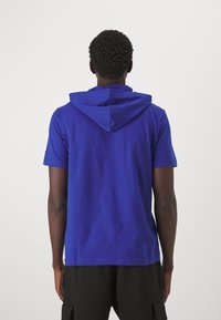 Niebieska bluza z krótkim rękawem z miękkiej bawełny, o luźnym kroju i dopasowanym kapturze z tyłu. Brak widocznych wzorów i elementów metalowych.