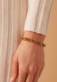 Gouden schakelarmband met ingewikkelde gedraaide en gladde secties, gedragen aan een hand, met een zacht textuur, lichtgekleurde gebreide trui.
