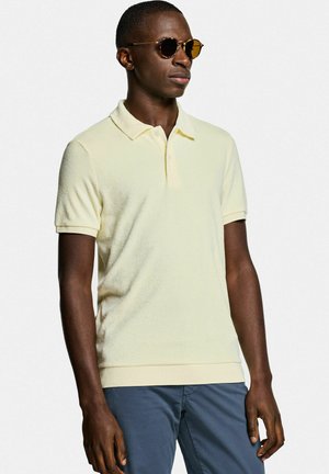 Homme portant un polo jaune à manches courtes, un pantalon bleu et des lunettes de soleil rondes marron, debout devant un fond clair uni.