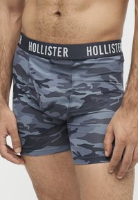 Homme portant un boxer à motif camouflage bleu et gris avec une ceinture noire marquée Hollister.