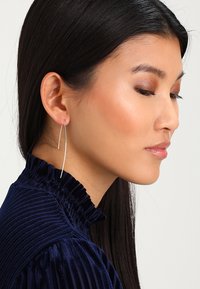Siva produžena naušnica na jednoj ušnoj resici, s elegantnim, minimalističkim dizajnom. Model ima tamnu kosu i nosi tamnoplavu bluzu s teksturom.