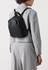 Sac à dos en cuir noir avec des accents de fermeture éclair dorés, de forme rectangulaire, surface texturée, porté sur une chemise à carreaux, avec des sangles réglables.