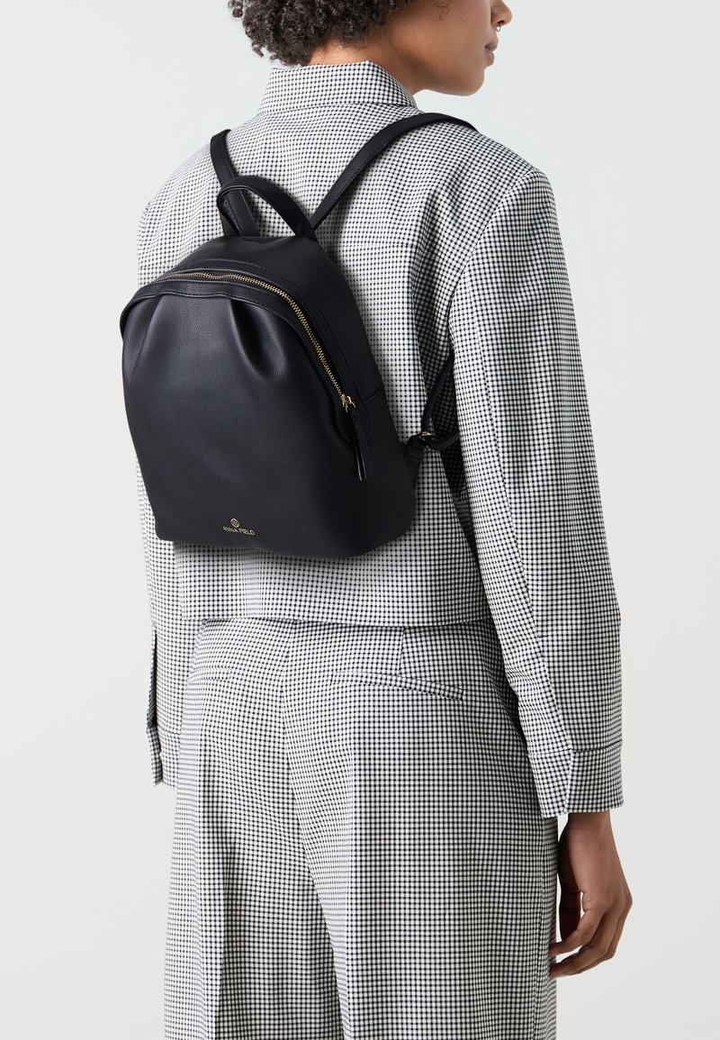 Sac à dos en cuir noir avec des accents de fermeture éclair dorés, de forme rectangulaire, surface texturée, porté sur une chemise à carreaux, avec des sangles réglables.