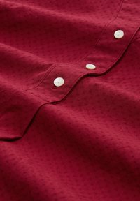 Bordeauxrotes Hemd mit strukturiertem Muster, weißen Knöpfen und klassischem Kragen. Der Stoff wirkt weich und leicht.