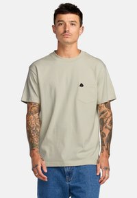 T-shirt verde in cotone con maniche corte, scollo rotondo e taschino sul petto con un piccolo logo nero. Indossato con jeans blu.