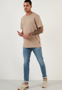Tricou de culoare maro deschis, cu mâneci scurte și gât rotund, combinat cu blugi slim-fit de culoare albastru deschis și teniși bej. Material neted, design simplu.