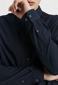 Granatowa koszula zapinana na guziki z długimi rękawami, gładką fakturą oraz dwoma guzikami na każdym mankiecie. Widoczny charakterystyczny kołnierz i przednia listwa guzikowa.