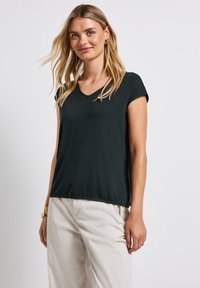 Temně zelený top s krátkým rukávem a výstřihem do V, volnější střih a mírně nařasený lem. Vyrobeno z měkkého, lehkého materiálu.