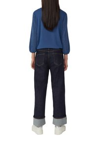Personne avec de longs cheveux sombres portant un haut à manches longues bleu, un jean foncé avec revers et des baskets compensées blanches, se tenant de dos.