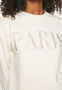 Felpa crema con testo embosso "PARIS" in un colore beige chiaro. Texture morbida, scollatura rotonda e maniche lunghe sono visibili.