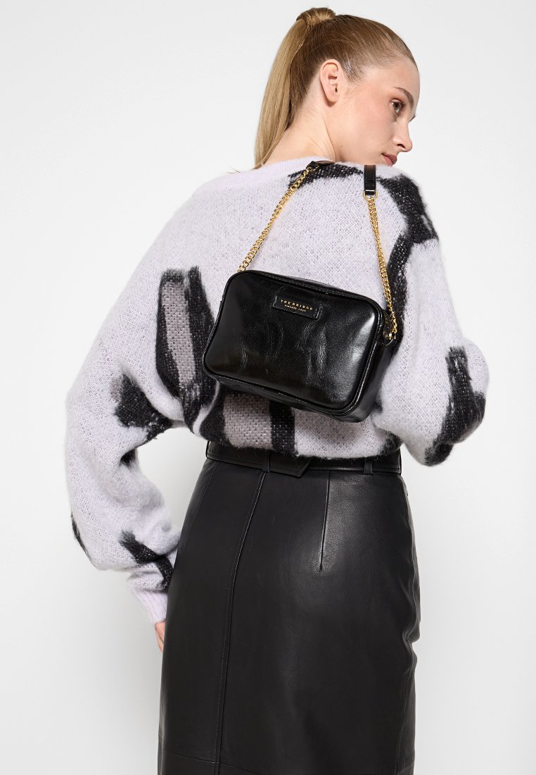 Borsa a tracolla in pelle nera con forma rettangolare, tracolla a catena dorata e superficie liscia. Indossata sopra un maglione viola chiaro e una gonna in pelle.