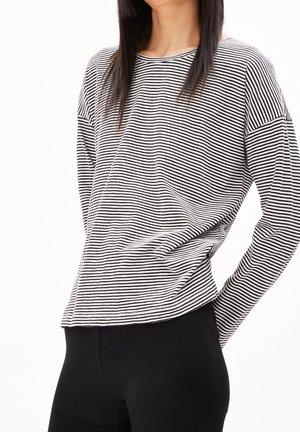 Femme portant un haut à manches longues rayé horizontal noir et blanc et un pantalon noir, posant avec une main sur la hanche.