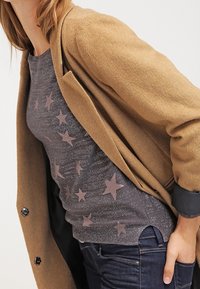 Tan ullblandad kashmirtröja över en grå topp med metalliska stjärnmönster. Mörka denimjeans. Toppen har en rund hals och åtsittande ärmar.