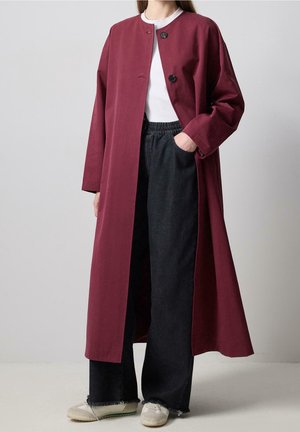 Person trägt langen burgunderroten Mantel mit schwarzen Knöpfen über weißem Oberteil, schwarze Hosen mit weitem Bein und weiße Turnschuhe, steht mit einer Hand in der Tasche.