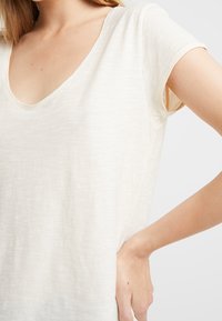 Svetle béžové tričko s jemnou texturovanou úpravou, s hlubokým V-výstřihem a krátkými rukávy. Design zdůrazňuje uvolněný střih.