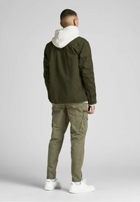 Man som bär en olivgrön jacka över en vit hoodie, ljusbruna cargobyxor och vita sneakers, stående och vänd bort från kameran mot en grå bakgrund.