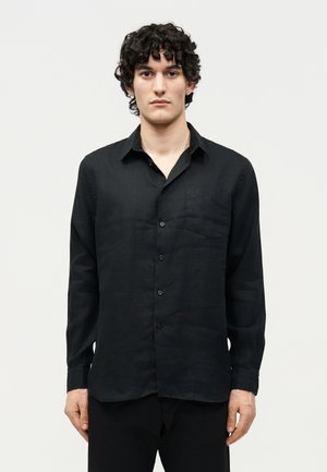 Νέος άνδρας με σγουρά σκούρα μαλλιά, που φοράει μακρυμάνικο μαύρο πουκάμισο με κουμπιά και μαύρο παντελόνι, μπροστά σε απλό λευκό φόντο.